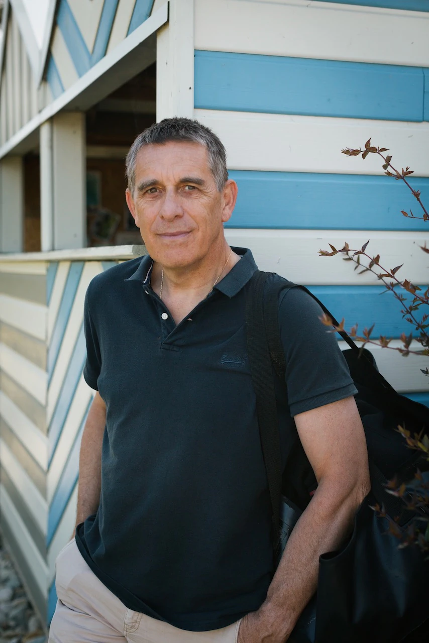 Portrait en gros plan d'un homme, une cinquantaine d'années, vêtu d'un t-shirt noir et adossé à un cabanon bleu et blanc